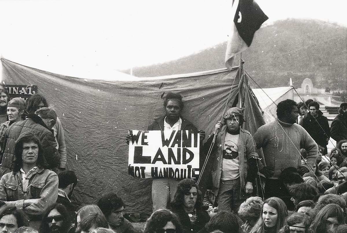 A man stands in front of a tent holding up a sign saying ‘We want land not handouts’. He is surrounded by a dozen or so other protesters.
