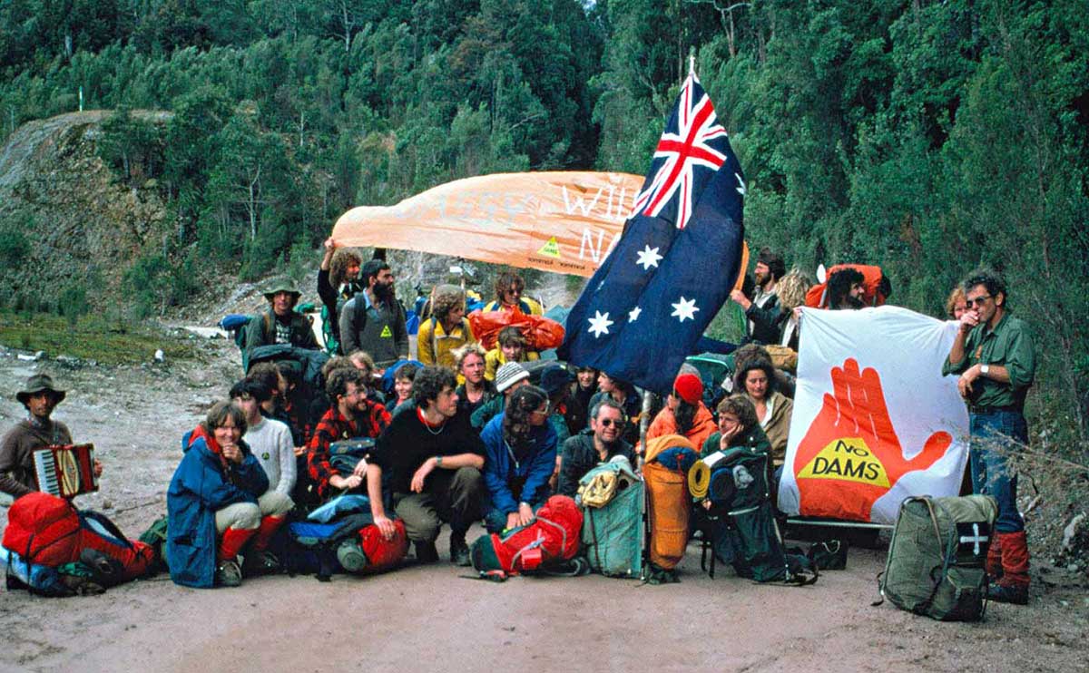 A group of protesters with hiking gear squatting or standing on a dirt road. They are holding the Australian flag and various banners, one that reads 'NO DAMS'.