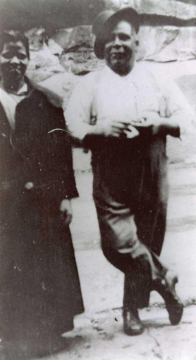Aboriginal man, smiling, stands facing the camera, a hat set at jaunty angle on his head. An Aboriginal woman stands to his right.