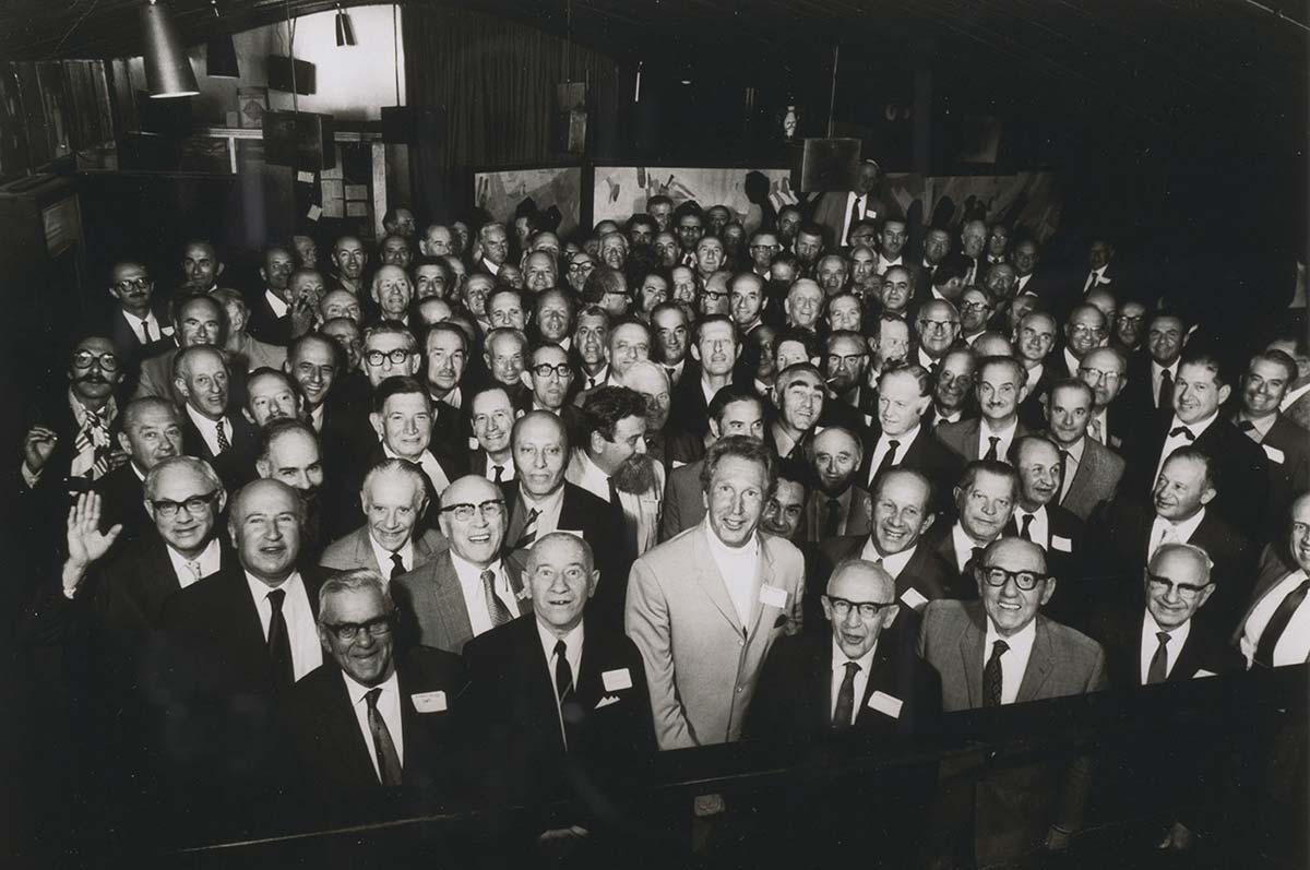 Interior photo of tightly packed group of about 70 men in their 50s and 60s smiling at the camera.