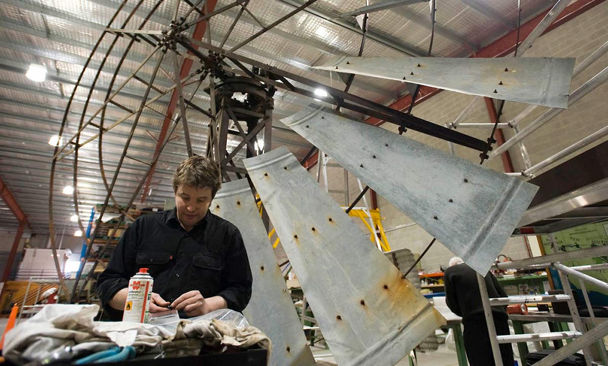 A man stands at a workbench, holding a small piece of metal. Behind him towers a partially re-assembled windmill.