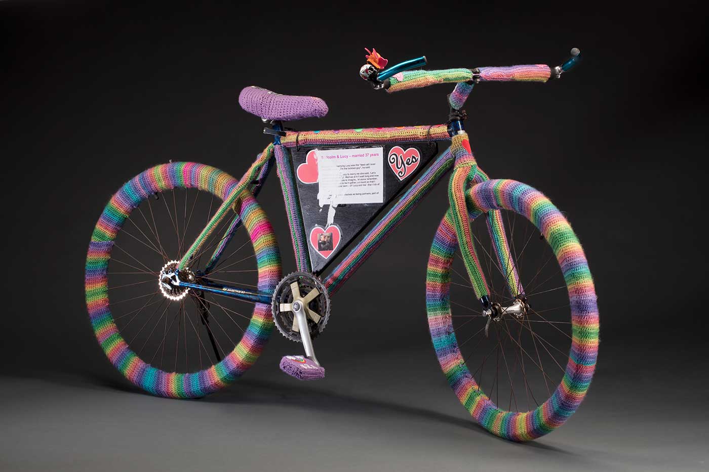 A bicycle covered with woollen crochet that consist of multi-coloured striped wheels, a purple seat and two purple peddles.