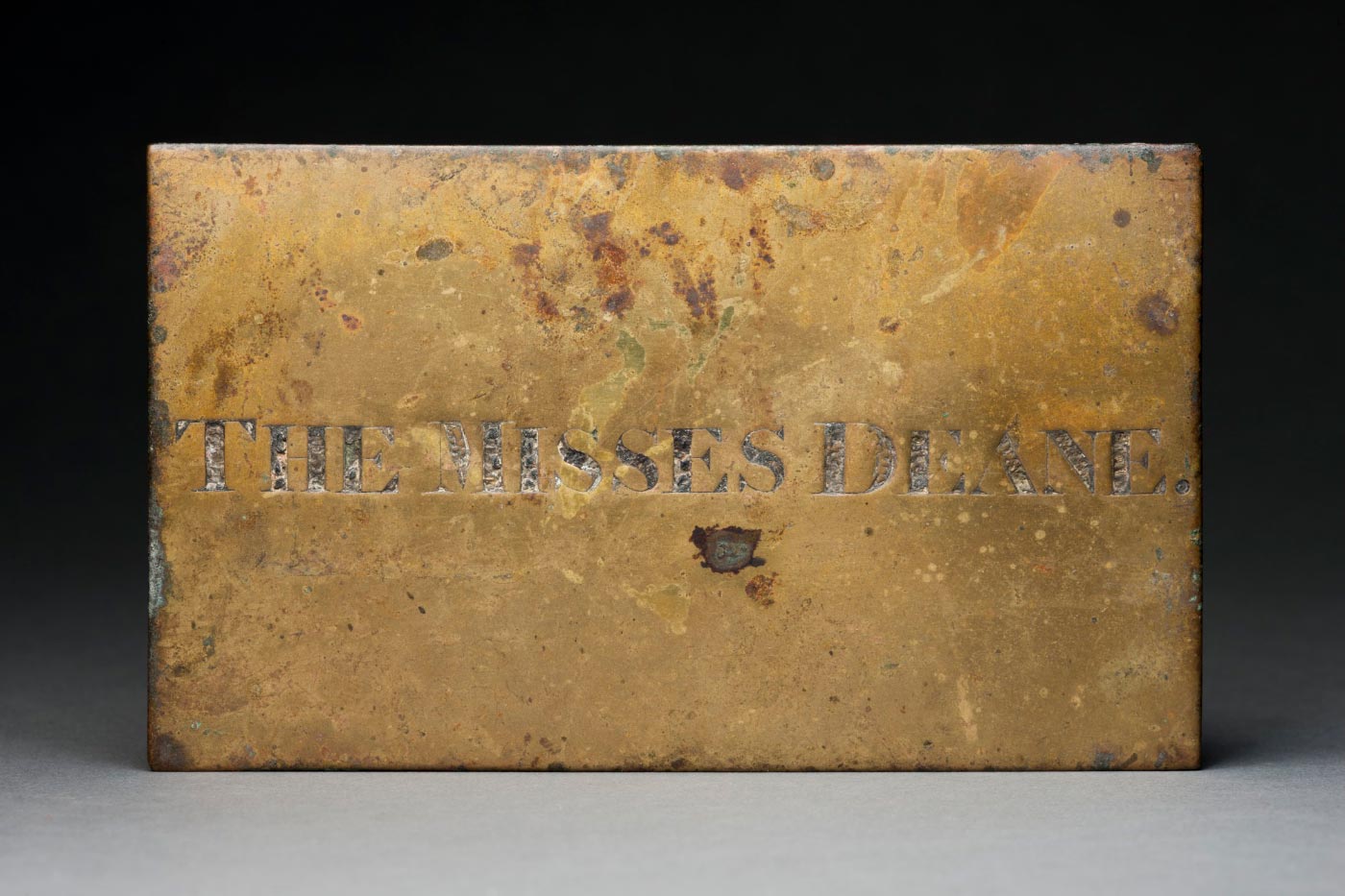 Brass name plate with the letters in capitals 'THE MISSES DEANE' engraved in the middle, showing signs of wear with several dents and metal clasps at the edges for hanging.