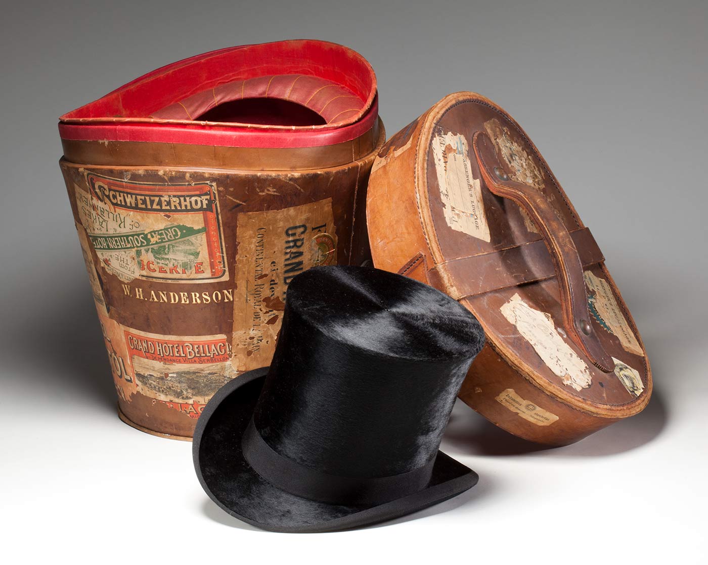 Black top hat made by J Woodrow & Sons, London, with hat box covered in travel stickers, featuring the inscription ‘W. H. ANDERSON’.
