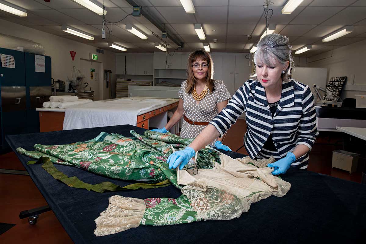Two women stand beside a laboratory bench, inspecting a green dress which is laid out on top. The woman on the right inspects the bodice.