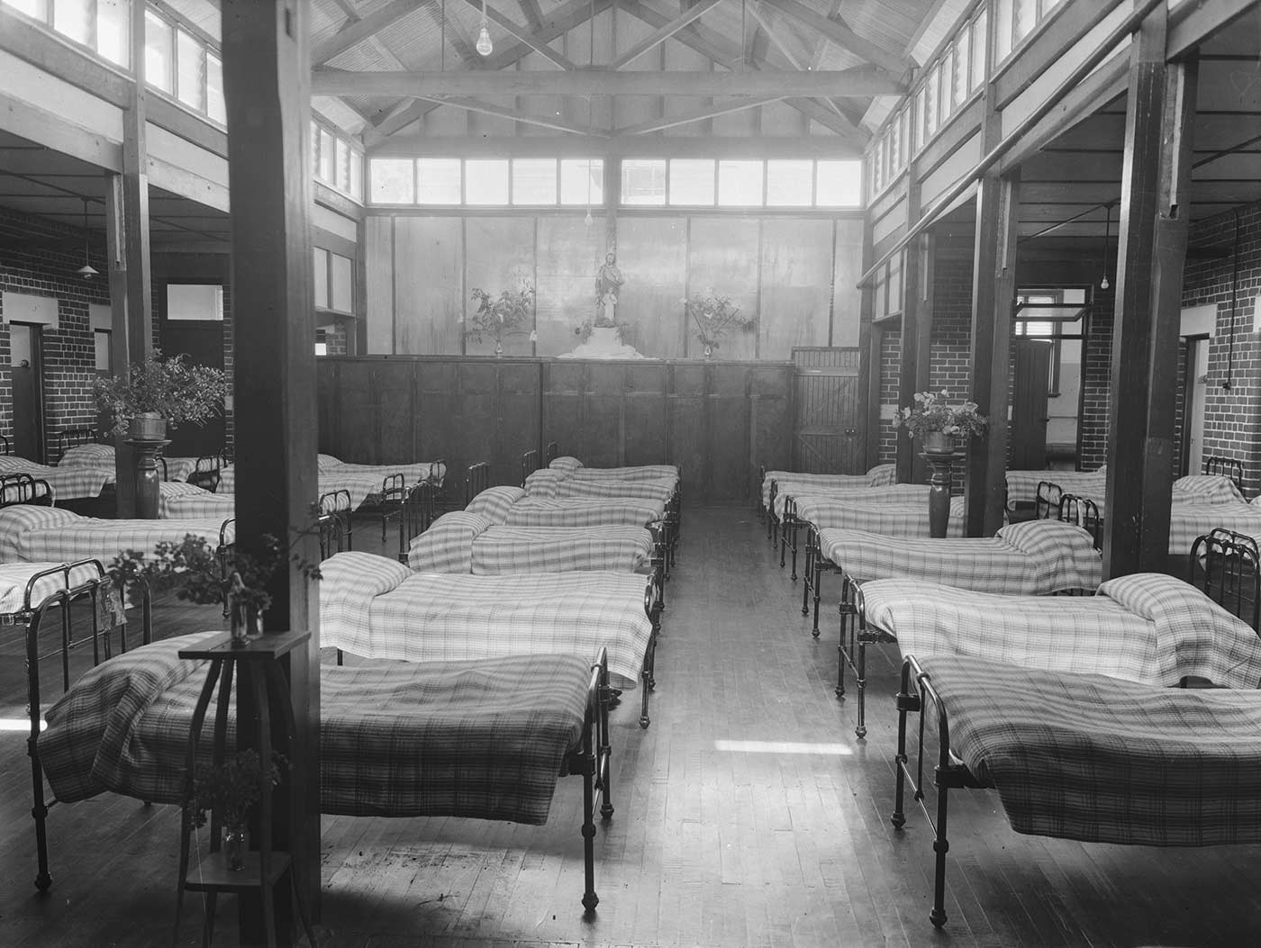 Black and white image showing a large room with high ceilings, filled with five rows of single, metal beds. Each is covered with a checked bedspread. Three small high tables stand in the room, with bases of flowers on top. Another two vases of flowers and a religious statue sit on top of cupboards at the far end.