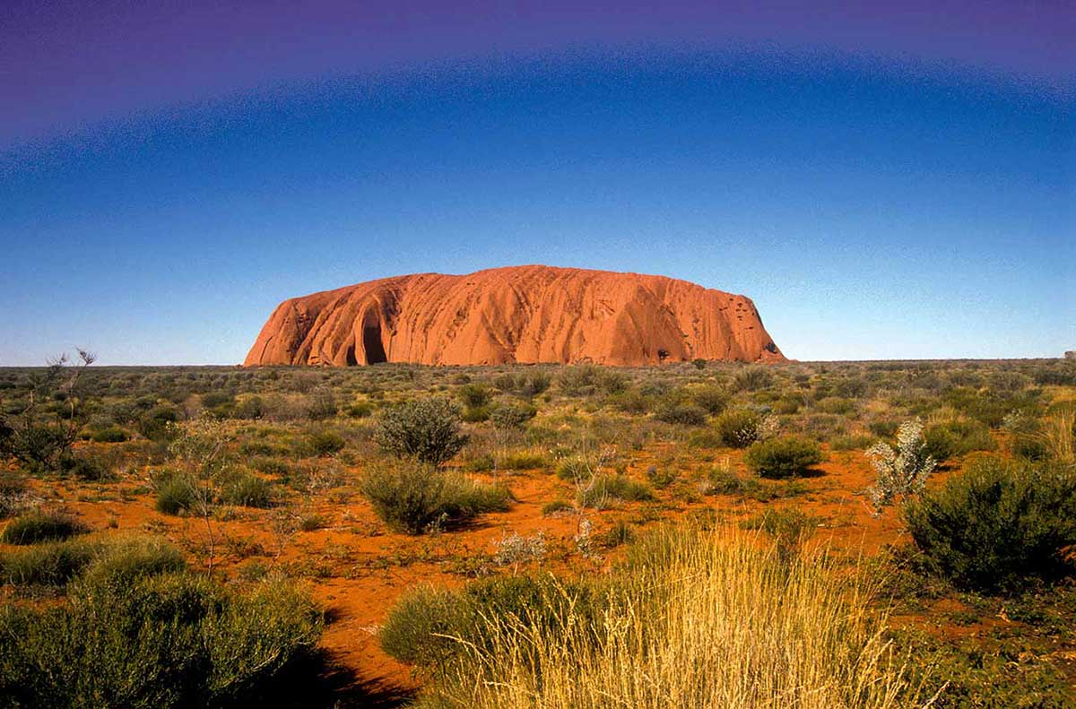 Panoramic view of a large red rock rising from a desert landscape..