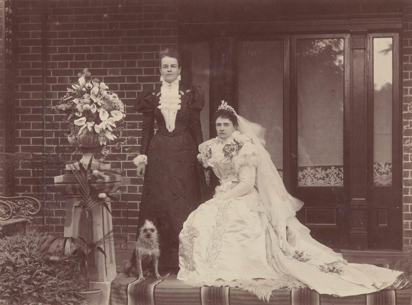 Black and white photo of a woman in a wedding dress and veil, seated. A woman in a dark dress stands at her left. A small terrier dog sits at her feet.