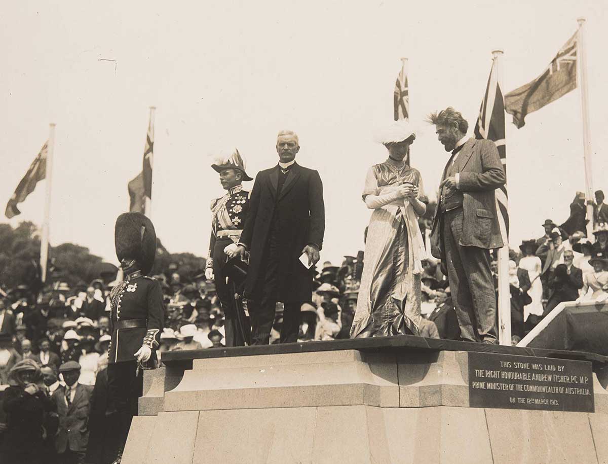 Black and white photograph of three men and a woman standing on a large stone monument as part of an official ceremony. A soldier in formal regalia stands nearby and a large crowd of spectators can be seen in the background.