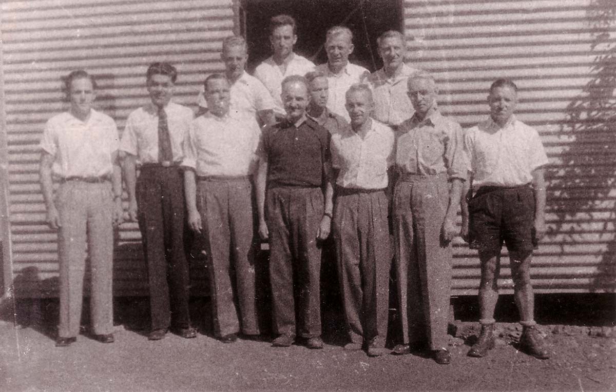 12 men, mostly wearing shirts and long pants, standing in front of a corrugated iron structure.