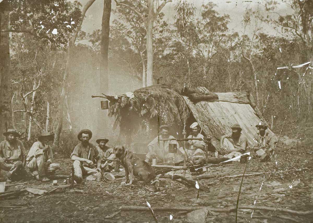 Black and white glass plate negative showing eight men sitting in front of a shelter in the bush. The A-frame shelter is made of bark and leaves and has a billy can hanging at the front. The men are gathered around a smoking fire, with another billy can hanging over the it. A dog sits at the front and centre of the image.