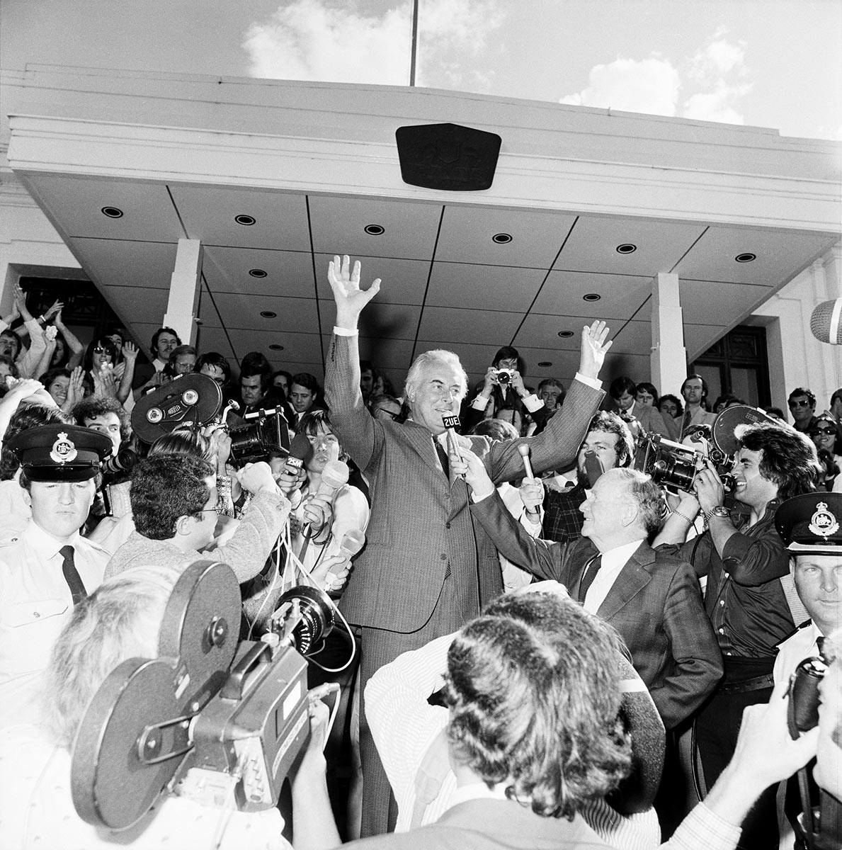 A man stands, addressing the media as he raises his hands to an applauding crowd.