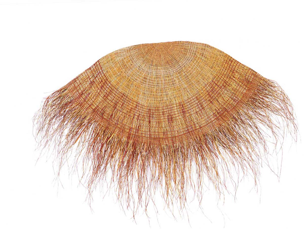 A mat made by an Indigenous Australian artist. The mat is round and is made from natural fibre. It is dyed in natural colours ie browns and ochres. Many loose strands hang from the edge of the mat.