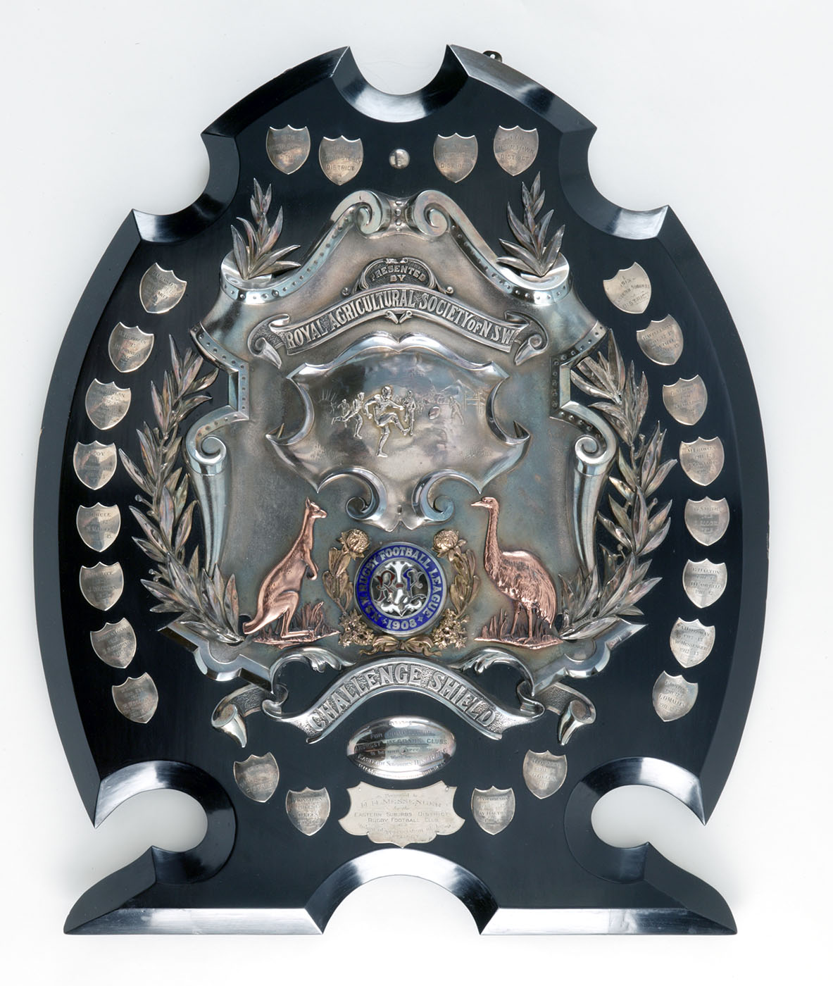 A sports shield with a football scene and coat of arms at the centre. Numerous small silver shield-shaped plaques are mounted around the edge of the trophy.
