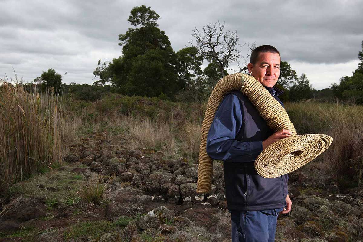 A man is holding a large vessel woven with a fibrous organic material. He is standing in a landscape featuring grassland, rocks and forest.