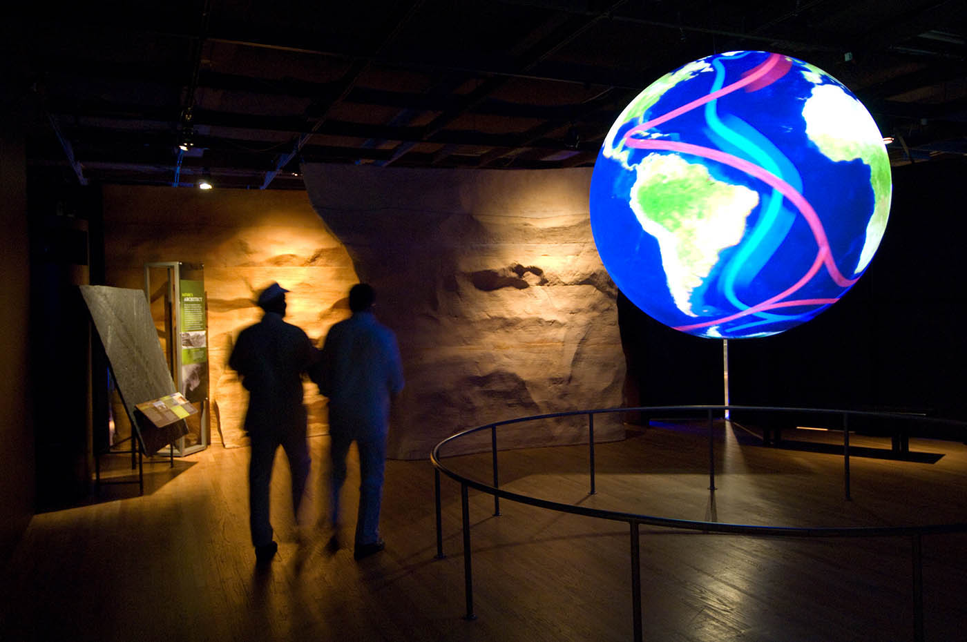 Two silhouetted visitors look at a large globe showing South America surrounded by a sea marked with multi-coloured water currents.