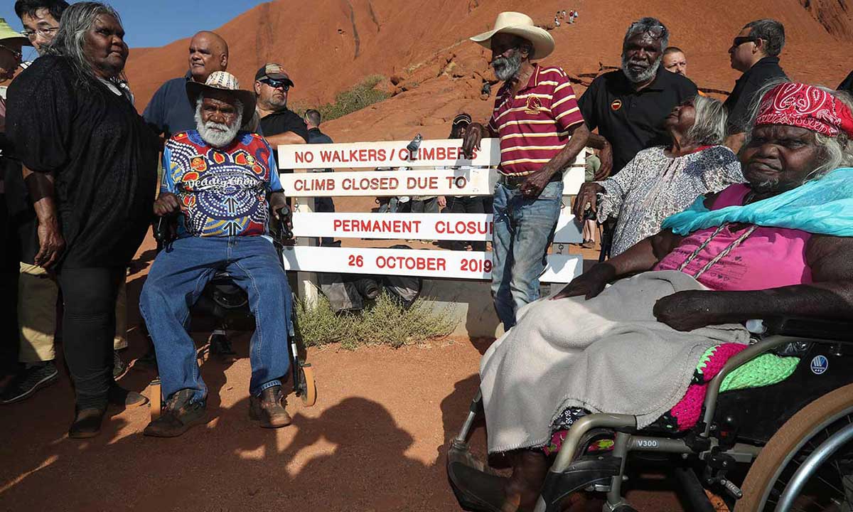 Colour photo of a group of people – some in wheelchairs – gathered around a sign that reads