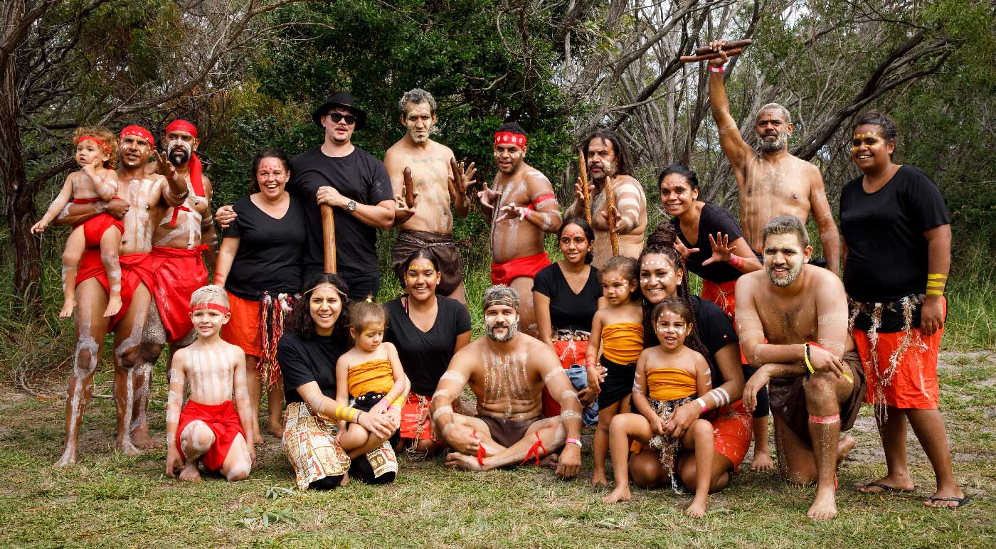 Portrait of a large group of men, women and children. They are dressed in traditional clothing and some are carrying wooden instruments.