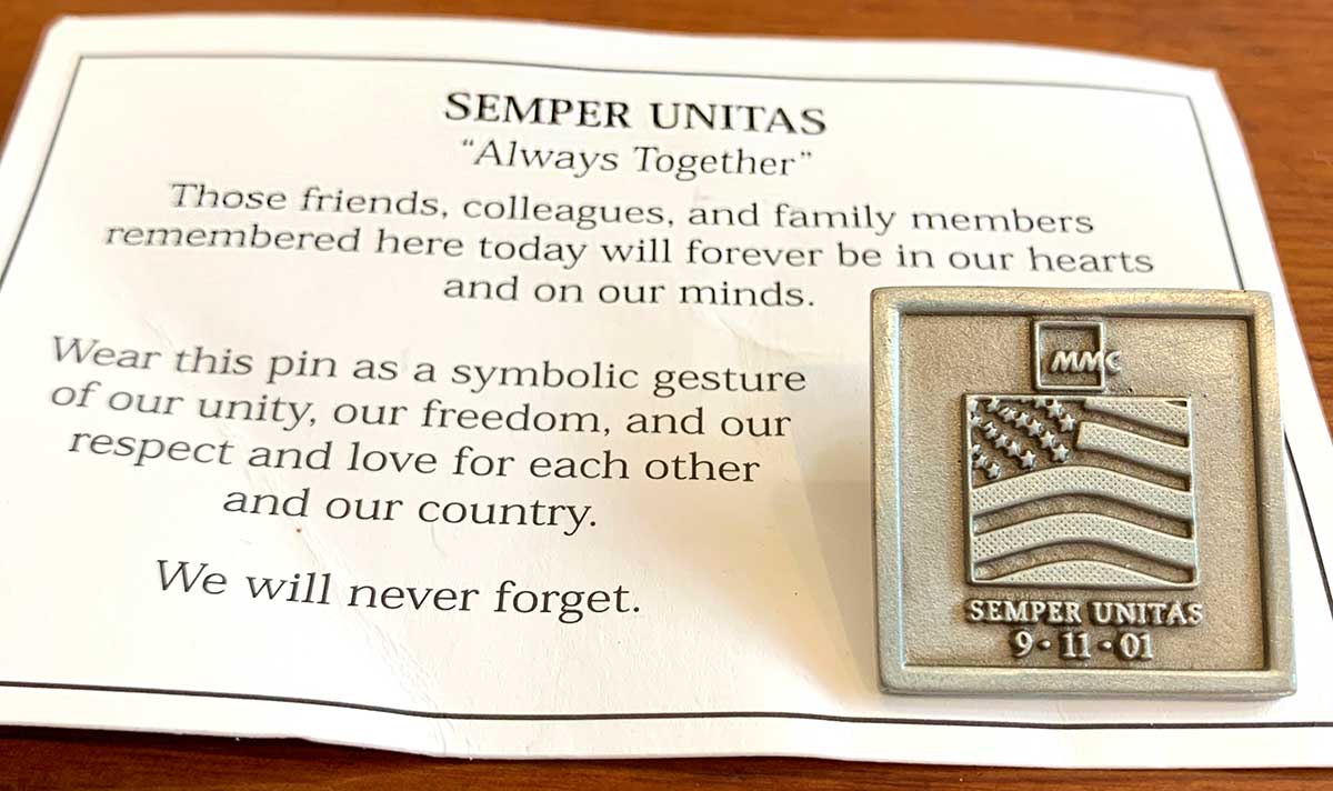 Small square silver pin with engraving of American flag, the letters MMC, and the words Semper Unitas 9-11-01. The pin is attached to a white card featuring black writing.