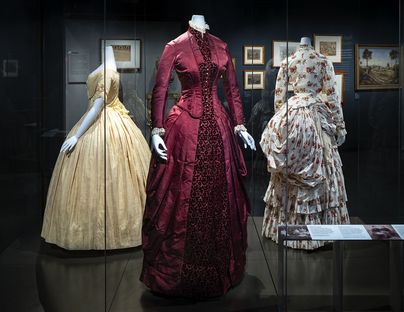 Three dresses on display in a large show case in the Landmarks gallery. The Burgundy dress is in the middle, to the left is golden cream silk dress and to the right is white dress with orange and red flowers.