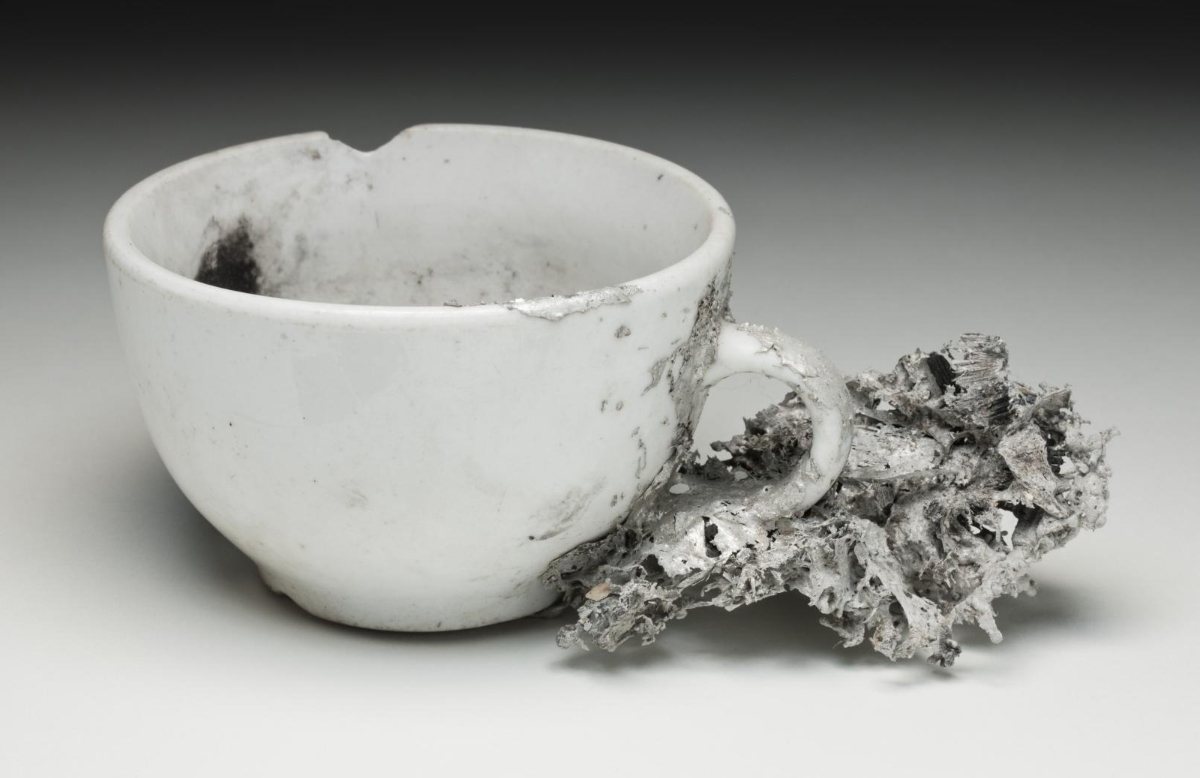 A photograph of a white teacup that has been damaged by fire. The cup has black soot marks, a chip in the lip of the cup and grey ash on the handle and outside of the cup.