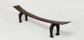Headrest kali Headrest made of wood, the ‘seat’ is slightly curved and supported by a pair of horseshoe-shaped legs. The ‘seat’ becomes wider from the middle towards both ends.