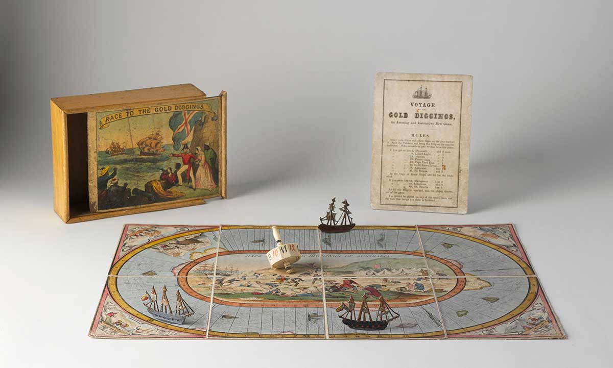 Studio photo of a game board with three pieces, an instruction manual and the box they came in. the box has a colourful illustration showing men holding a flag pointing at ships at sea. The pieces on the game board are ships and the board itself suggests that the game involves moving around an oval sea with an illustration of gold diggings in the centre.