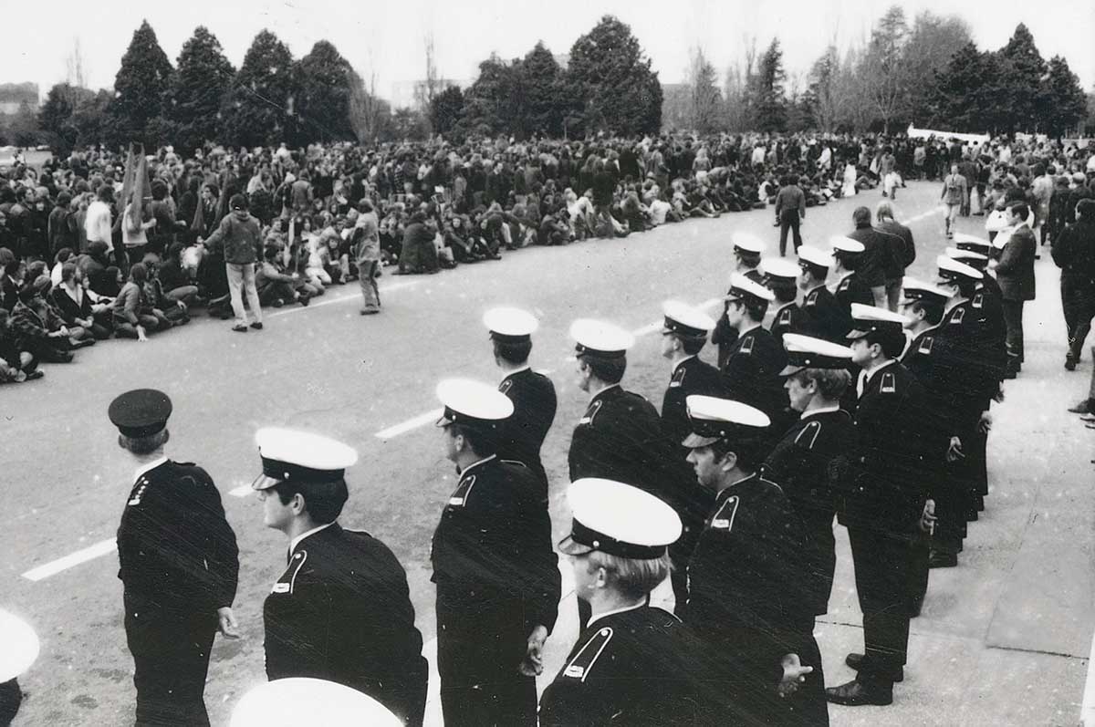 Two ranks of police line the street with a large crowd gathered on the other side.