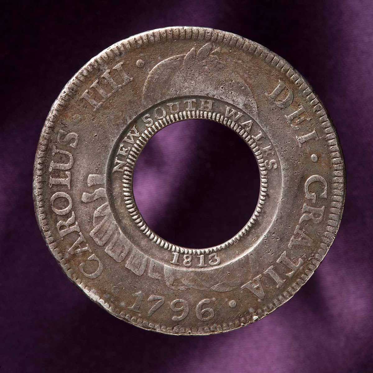 Circular coin with a hole at the centre. 'NEW SOUTH WALES 1813' is stamped around the centre ring. 'CAROLUS IIII DEI GRATIA 1796' is stamped around the outer rim.