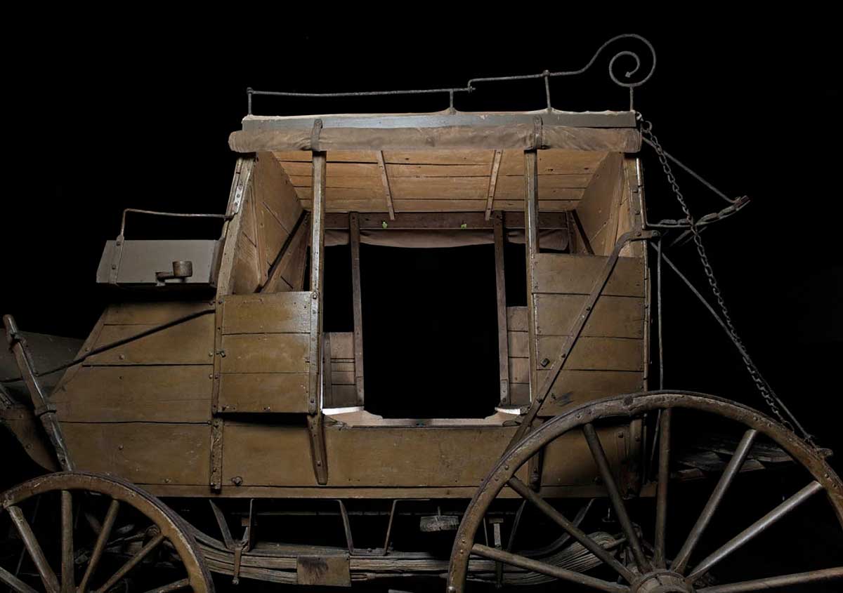 Wooden mail coach, painted brown, with canvas awnings and larger wheels at the rear.