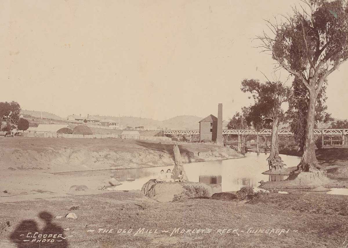 Old black and white photo of the mill in the distance surrounded by bare landscape and a few trees to one side. A river is in the foreground.