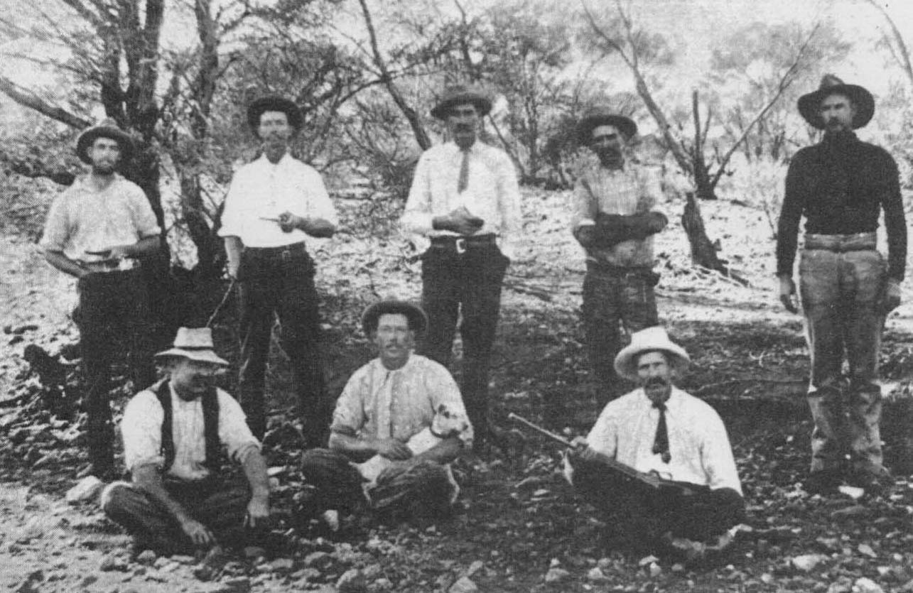 Black and white photo of eight men and a dog posing for a group photo in bushland.