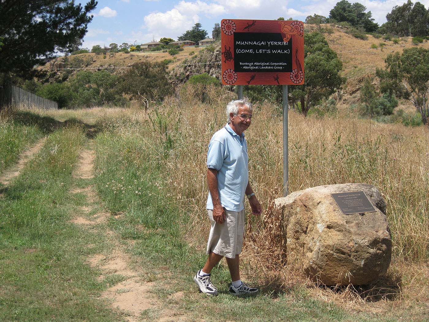 Eric Bell standing beside a dirt track, with a hill rising in the background. He is standing next to large rock with a commemorative plaque and a sign that reads 'MUNNAGAI YERRIBI (COME, LET'S WALK). Buranya Aboriginal Corporation - Aboriginal Landcare Group -'