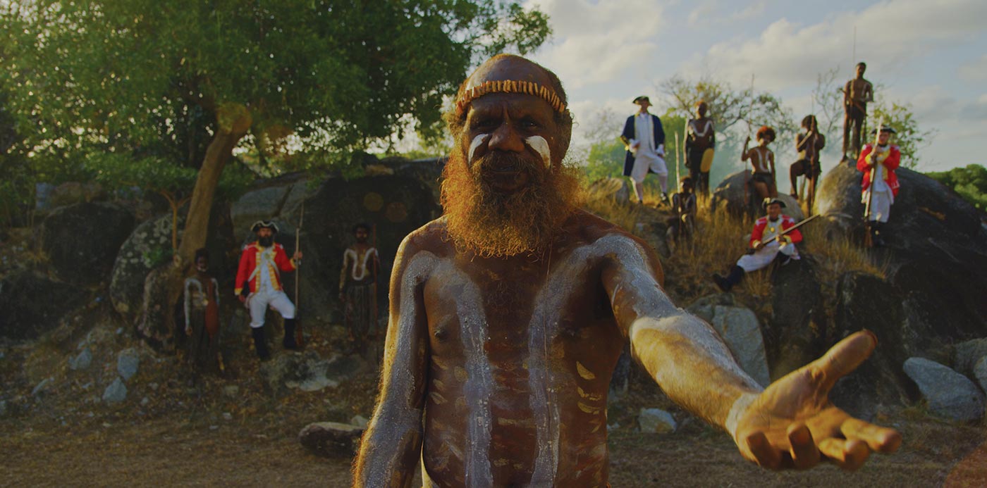 A bearded man with white paint on his face, chest and arms extends his open left palm to the camera. Behind him, soldiers and Indigenous people rest on large rocks.