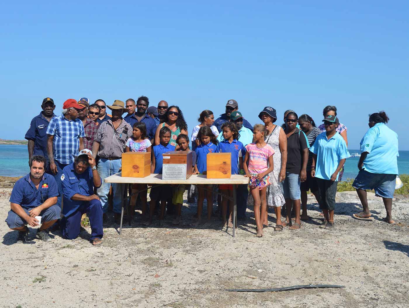 Photo of a large group of people standing around a table with boxes. In the background is a large river.