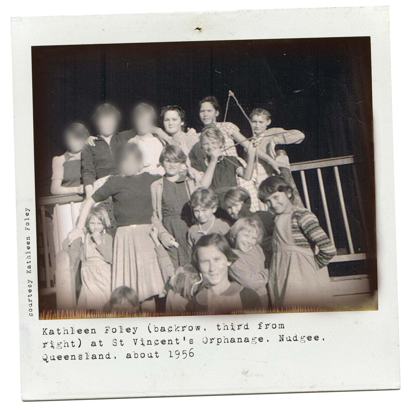 Polaroid photograph showing a group of girls standing at the front of a timber verandah rail. One of the girls holds a small bow and arrow. Some of the older girls' faces have been blurred. Typewritten text at the bottom reads 'Kathleen Foley (back row, third from right) at St Vincent's Orphanage, Nudgee, Queensland, about 1956'. 'Courtesy Kathleen Foley' is typed along the left side.