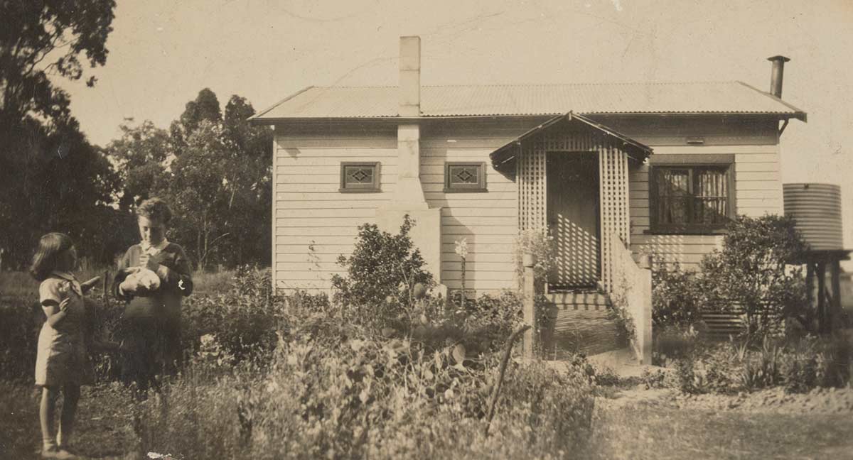 A black and white photograph of a young girl and boy standing in a garden in front of a weatherboard cottage.