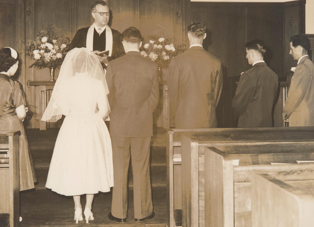 A black and white wedding photograph of seven people in a church, five men and two women. Standing on the left are two women, one of whom is the bride. To the right of her are four men. They are all facing away from the camera, standing in front of a man who appears to be a priest.