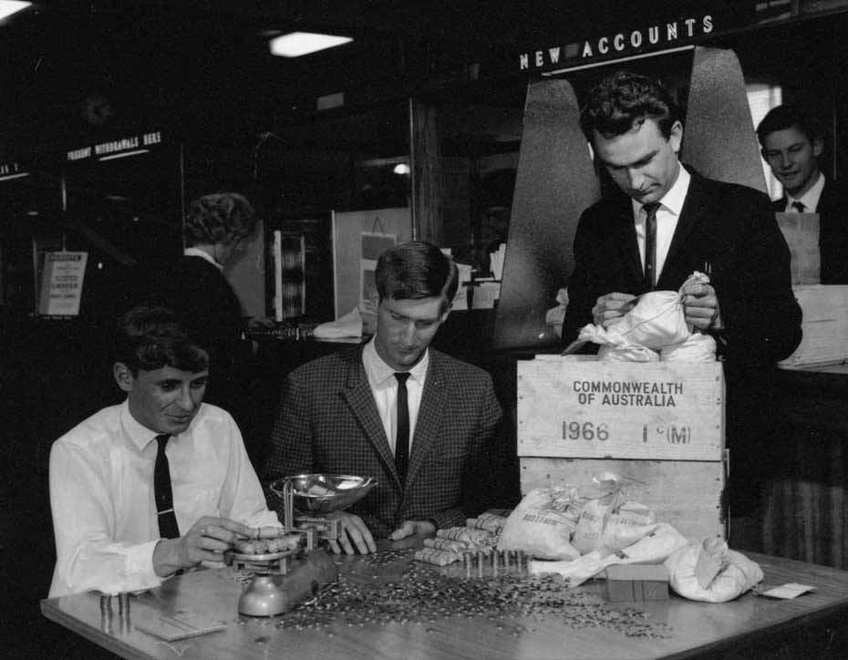 Black and white photograph of a group of men at a table counting, weighing and bagging coins for the Commonwealth of Australia.