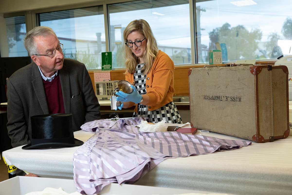 A man and woman view objects on a table, including a top hat, purple dress and suitcase.