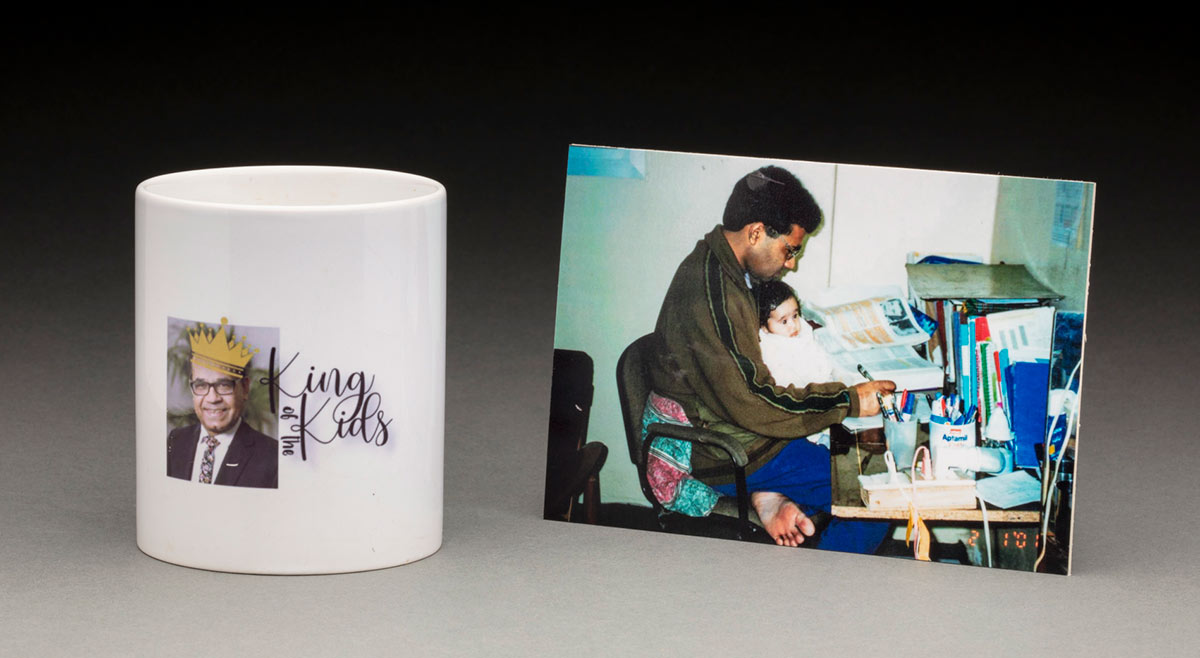 A white ceramic coffee cup with a colour portrait of a man on one side. Printed over the man's head is a yellow crown, and to the right is black text that reads 'King / of the / Kids'. A colour photograph of a man and child sits adjacent.