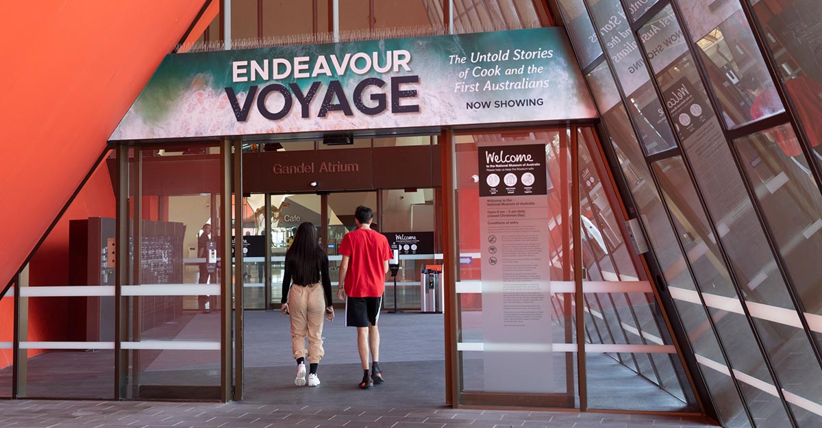 Two visitors walking through a large entrance with a promotional banner for the Endeavour Voyage exhibition above.