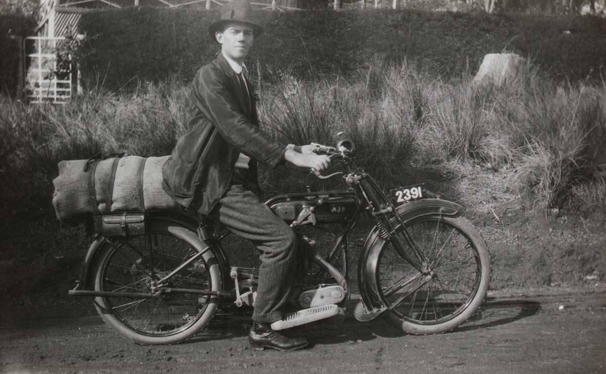 A black and white photograph of a man sitting stationary on a motorcycle with cargo strapped to the back. The number plate reads '2391'.