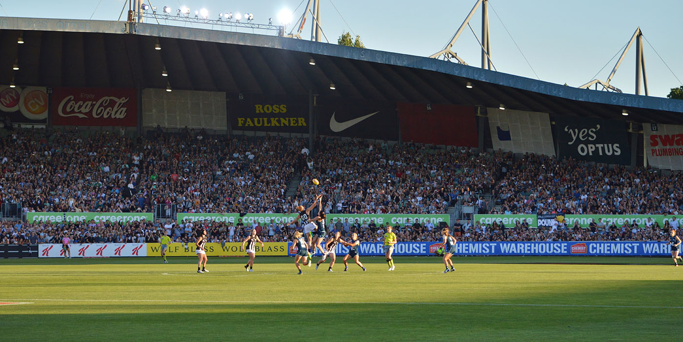 A photograph of an AFL game. Two players are competing for the ball whilst other players huddle around. Behind the players is stadium seating with lots of people watching the AFLW game.