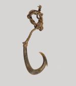 Fishhook makau paweo Fishhook made of mother-of-pearl with a curved shank and a barb located on the outer side of the hook, directly below the point. A short brown fishing line is attached.