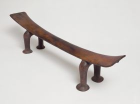 Headrest kali Headrest made of wood, the ‘seat’ is slightly curved and supported by a pair of horseshoe-shaped legs. The ‘seat’ becomes wider from the middle towards both ends.