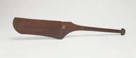 304.jpg Dance paddle made of wood with an almost rectangular and rounded blade (that is missing on one side) and a semi-circular knob at the end of the handle.