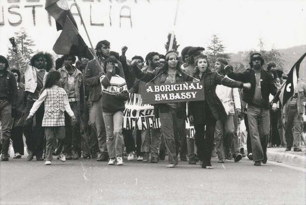A group of people walk along a road, holding flags and banners. A woman at the front has a placard around her neck reading ‘Aboriginal Embassy’.