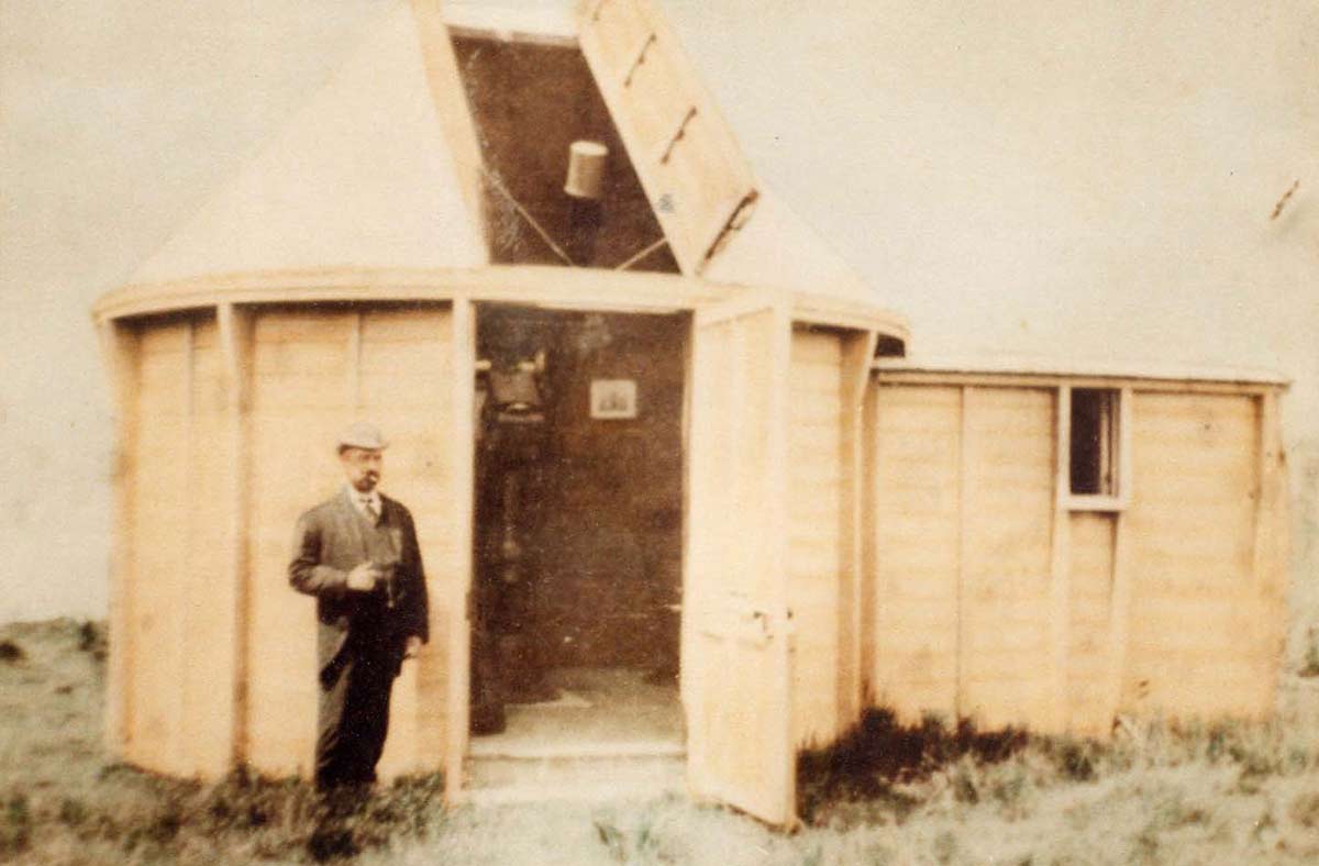 A man wearing a suit and hat stands beside a timber observatory building. The door and the roof of the circular structure are open, revealing a telescope inside.