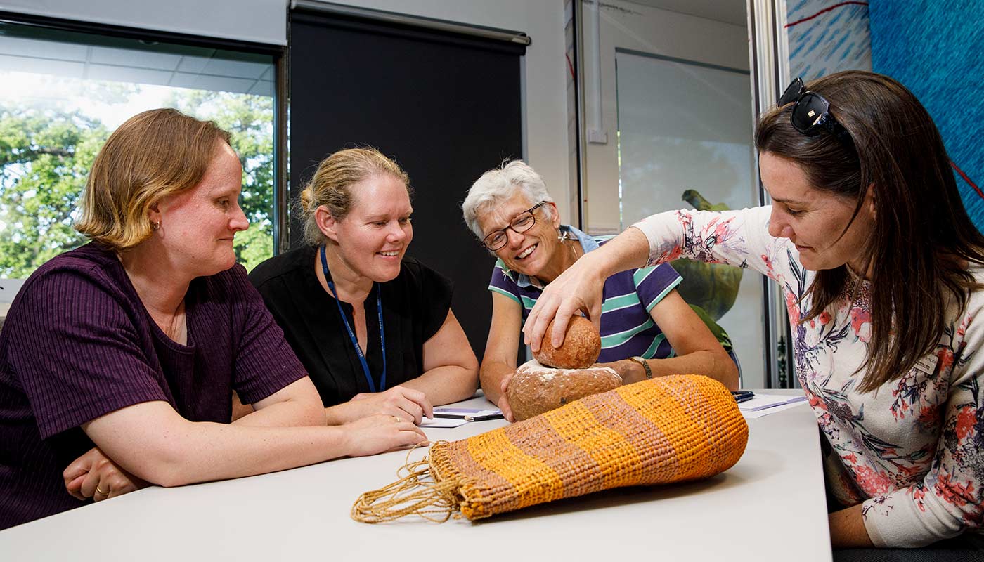 Three women sitting at a table inspecting stone tools and a woven bag.